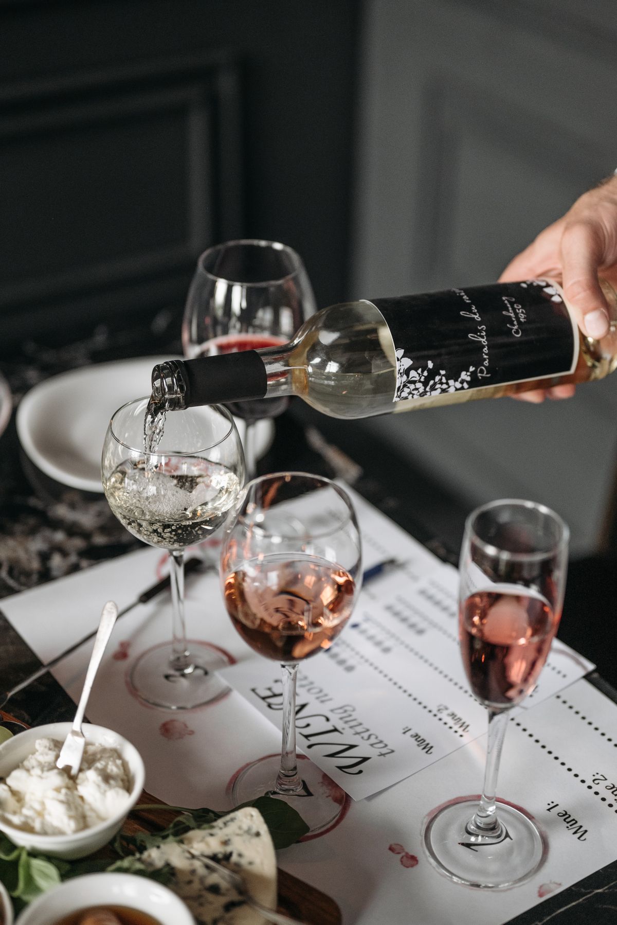 White wine being poured into a glass at a home wine tasting, with cheese and tasting sheets on the table